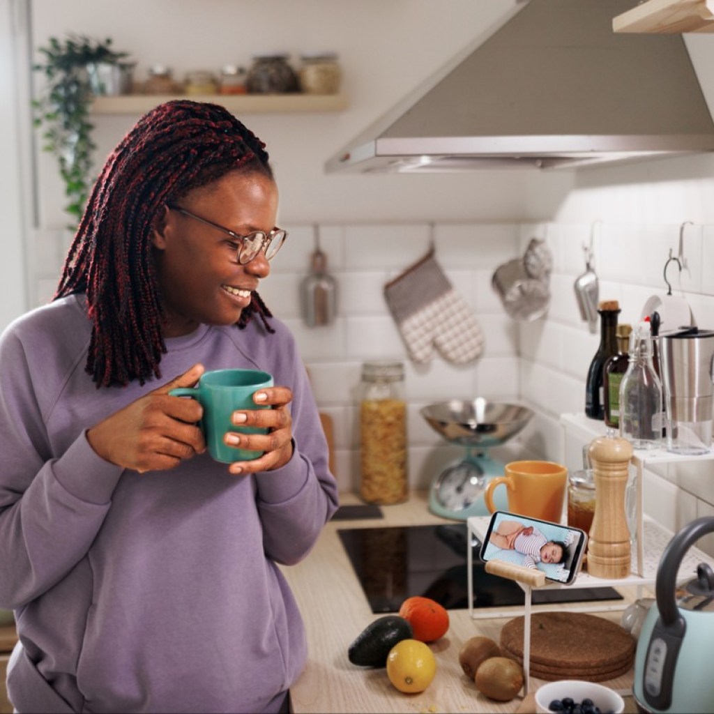 Smiling woman having coffee in kitchen using phone to monitor her sleeping baby